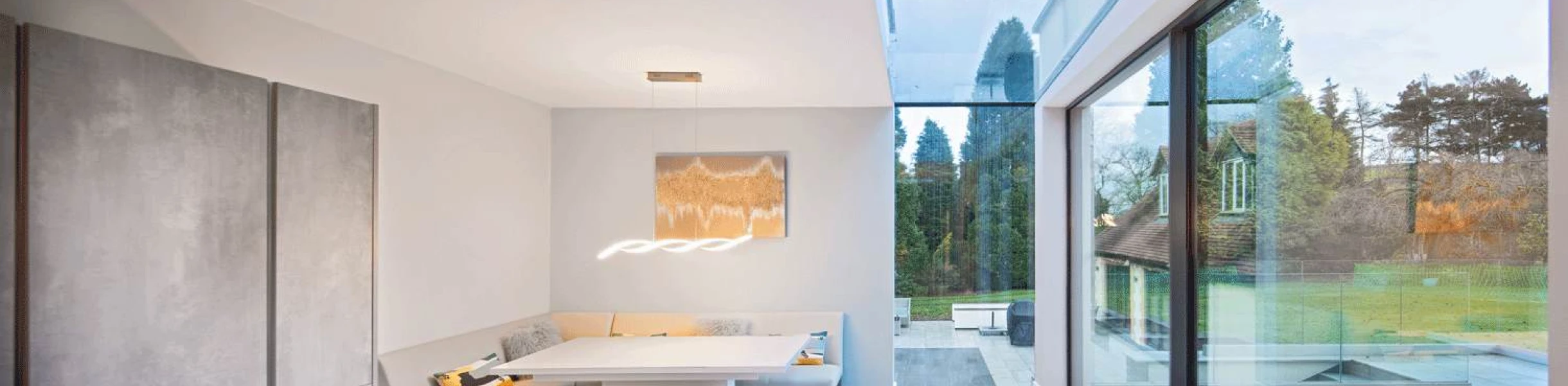 Intérieur moderne d'une salle à manger avec une table en bois, un coin repas confortable, de grandes fenêtres offrant une vue sur un jardin verdoyant, et un éclairage suspendu contemporain.