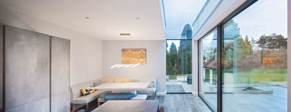 Helle moderne Küche mit einem Essbereich in einer Nische und großen Fenstern, die den Blick auf einen Garten freigeben. Ein minimalistischer Tisch und eine stilvolle Beleuchtung ergänzen das Design.
