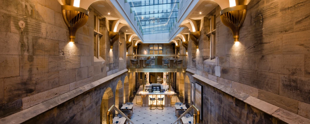 A grand stairway leads down into a stylish interior space featuring stone walls and warm lighting. A glass ceiling allows natural light to illuminate the area, which includes elegant tables and modern décor.