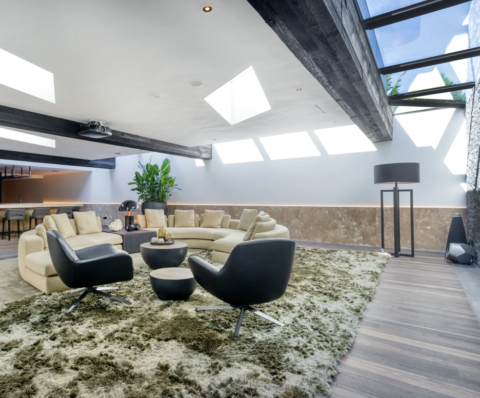 Sala moderna y acogedora con un sofá en forma de U, sillones negros y una alfombra suave. Ventanales en el techo permiten la entrada de luz natural. Al fondo, una cocina elegante y minimalista.