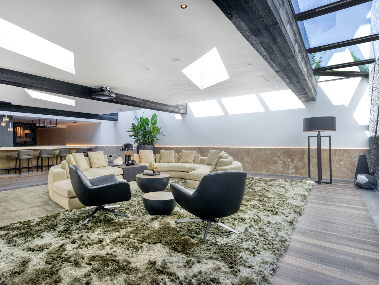 Sala moderna y acogedora con un sofá en forma de U, sillones negros y una alfombra suave. Ventanales en el techo permiten la entrada de luz natural. Al fondo, una cocina elegante y minimalista.