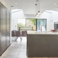 Modern kitchen with gray cabinetry, an island with a countertop, and a view of an outdoor dining area through large glass doors. Natural light floods the space from skylights and windows. Stylish pendant lights hang above the island.