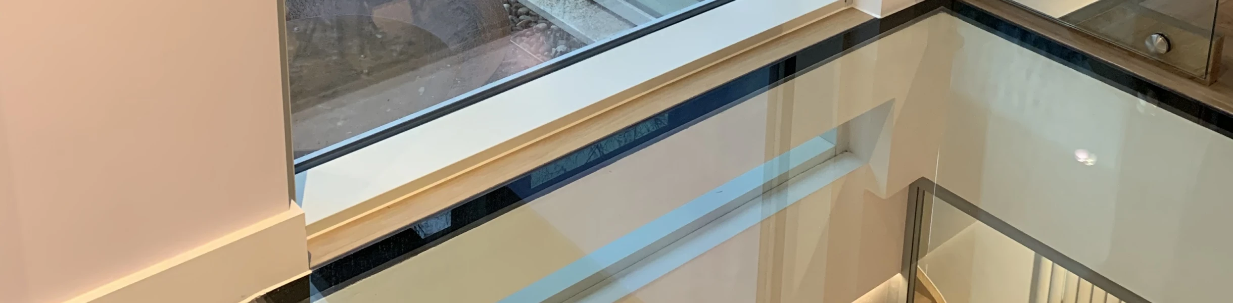 Large modern window with a view of a patio and garden. A reflection of a person is visible on the glass. The interior features a sleek design with a glass floor and grey tiles.
