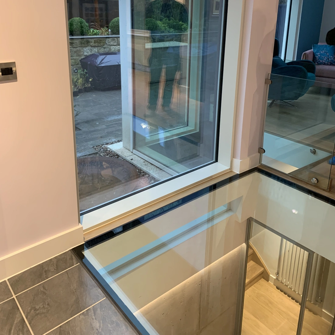 Großes Fenster zeigt die Außenseite eines modernen Hauses mit einem Spiegelbild der Person im Inneren. Der Boden ist aus Glas und gibt einen Blick auf die darunterliegende Ebene frei.