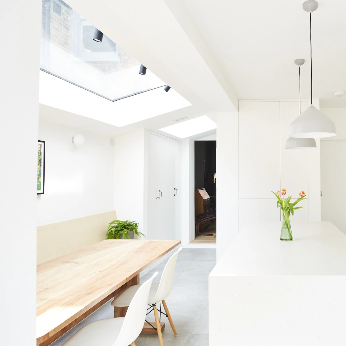 Cocina moderna y luminosa con paredes blancas, una isla central, electrodomésticos integrados y una mesa de madera. Flores en un jarrón adornan la isla.