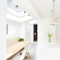 Cocina moderna y luminosa con paredes blancas, una isla central, electrodomésticos integrados y una mesa de madera. Flores en un jarrón adornan la isla.