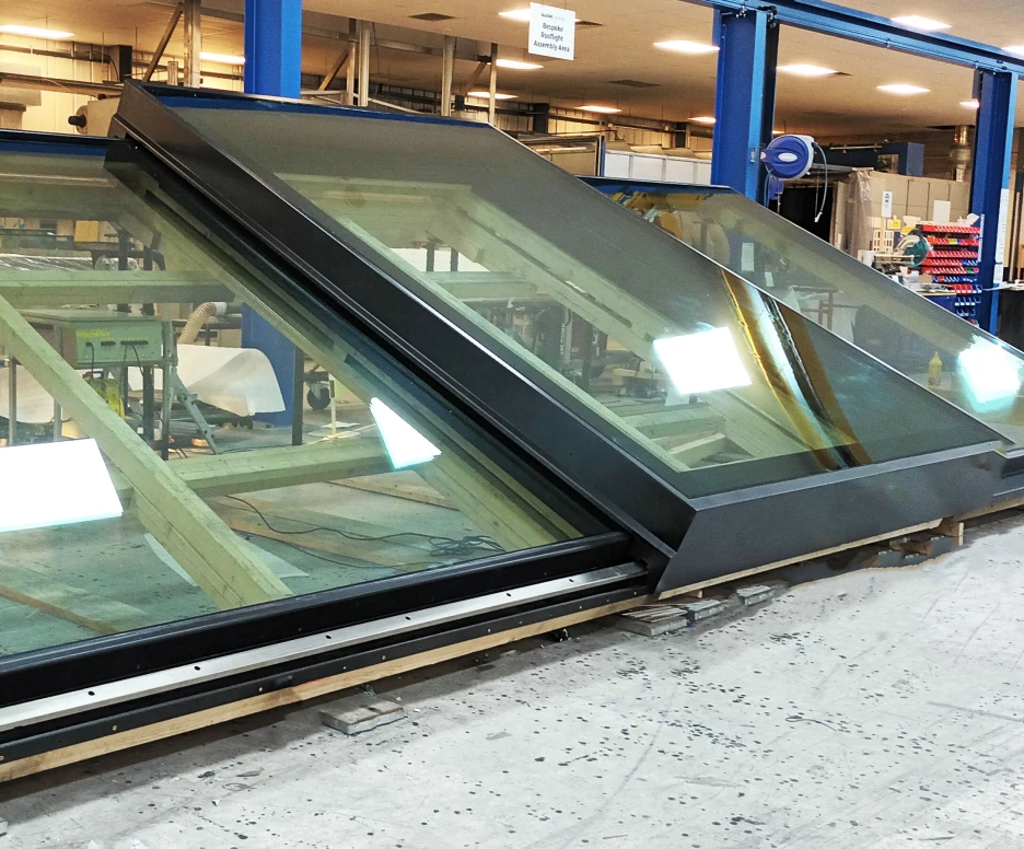 Large, angled glass skylights with black frames are positioned on wooden supports in a workshop. The floor is concrete, and various tools and materials are visible in the background.
