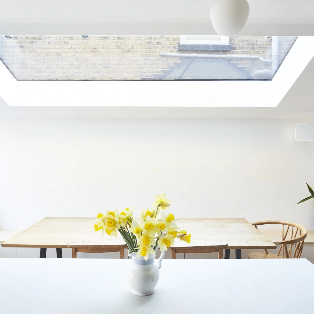 Une cuisine lumineuse avec une grande fenêtre de toit. Une table en bois est placée au centre, entourée de chaises. Un vase blanc contient des jonquilles jaunes sur un comptoir blanc. Une plante verte est visible à droite.