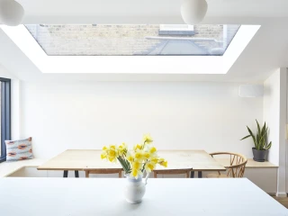 Bright, minimalist kitchen featuring a large skylight, wooden dining table with chairs, and a vase of yellow flowers on the counter. A potted plant adds a touch of greenery.