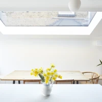 Une cuisine lumineuse avec une grande fenêtre de toit. Une table en bois est placée au centre, entourée de chaises. Un vase blanc contient des jonquilles jaunes sur un comptoir blanc. Une plante verte est visible à droite.