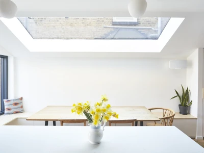 Interior de una cocina luminosa con una ventana de techo. En el centro, una mesa de madera rodeada de sillas y un jarrón con flores amarillas. Una planta verde se encuentra en la esquina y hay un cojín decorativo en el fondo.