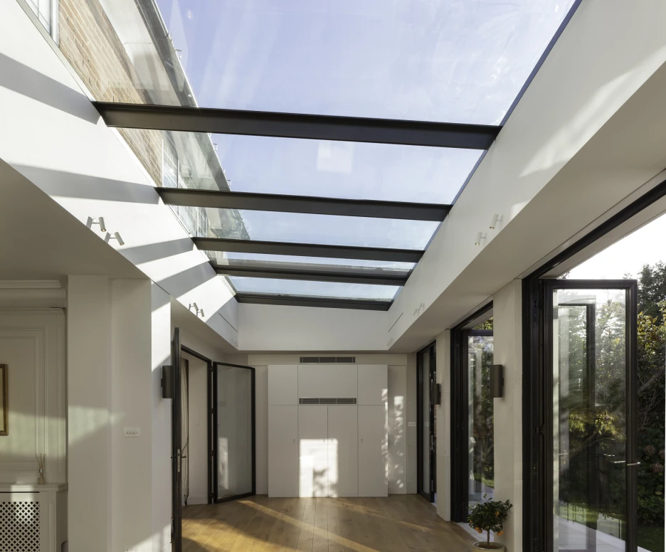 Interior de una habitación con gran entrada de luz natural a través de un techo de cristal. Suelo de madera clara y puertas de vidrio que se abren hacia el exterior. La decoración es minimalista con un toque moderno.
