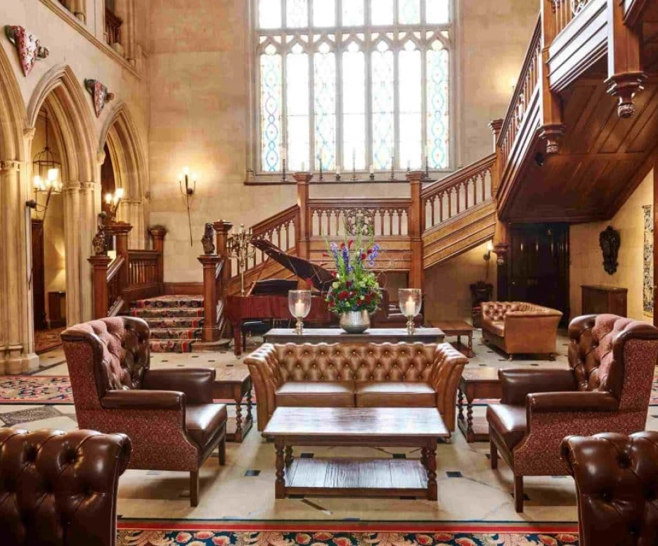Luxurious interior of a grand hall featuring a staircase, ornate wooden details, a grand piano, and elegantly upholstered seating. Natural light streams through stained glass windows, highlighting a decorative floral arrangement on a central table.