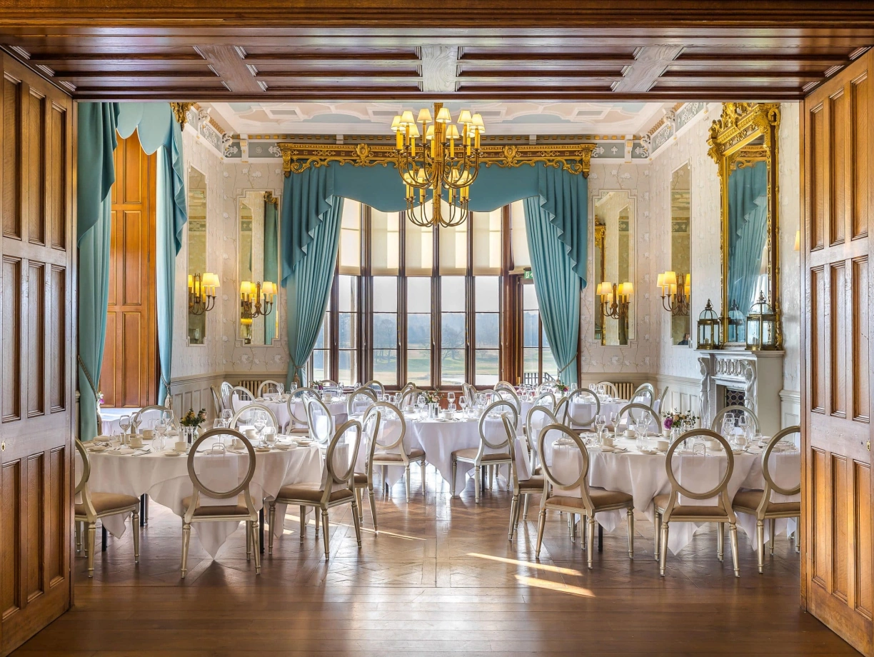 An elegant dining room with round tables set for a formal meal. The walls are adorned with intricate moldings, and large windows draped with teal curtains allow natural light to fill the space. Chandeliers hang from the ceiling, contributing to the sophisticated ambiance.