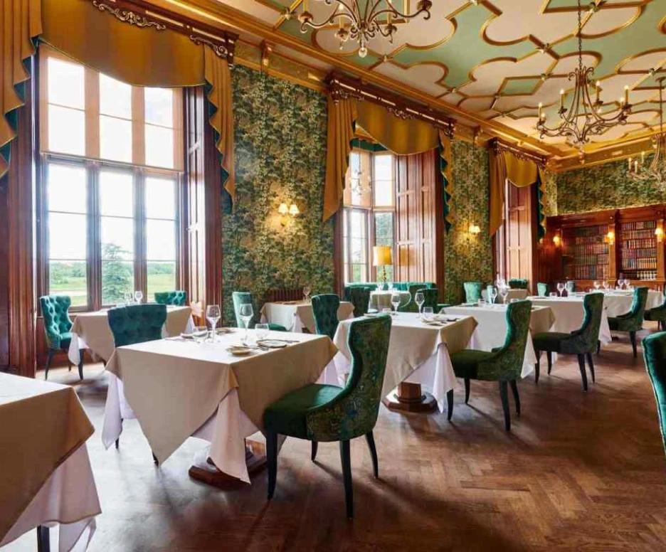 An elegant dining room featuring green floral wallpaper, large windows with golden drapes, and round tables covered with white tablecloths. Velvet green chairs surround the tables, while a bookshelf is visible in the background. Chandelier lights hang from the ceiling, adding to the luxurious ambiance.