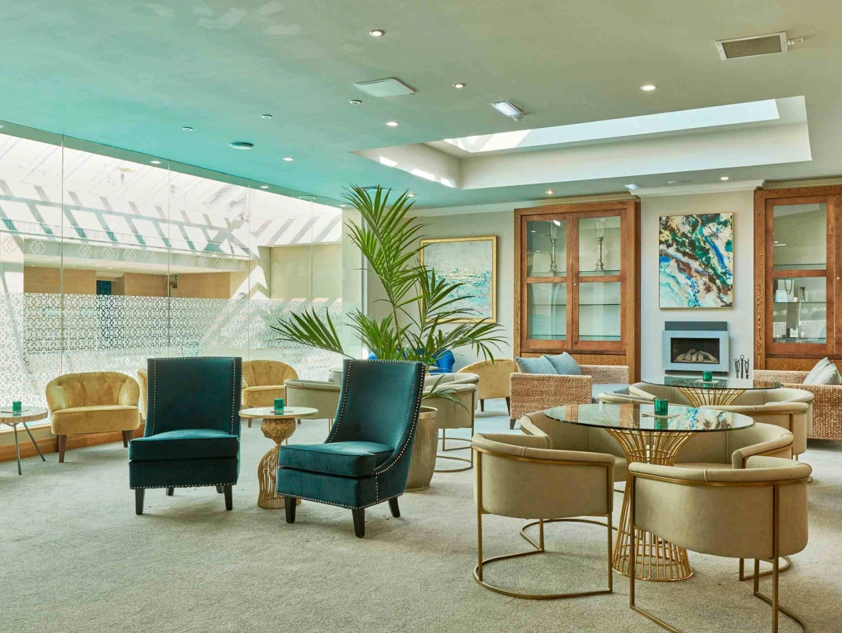 Un salon spacieux et lumineux avec des fauteuils confortables, des tables rondes et de grandes fenêtres laissant entrer la lumière naturelle. Des plantes décoratives ajoutent une touche de verdure à l’espace.
