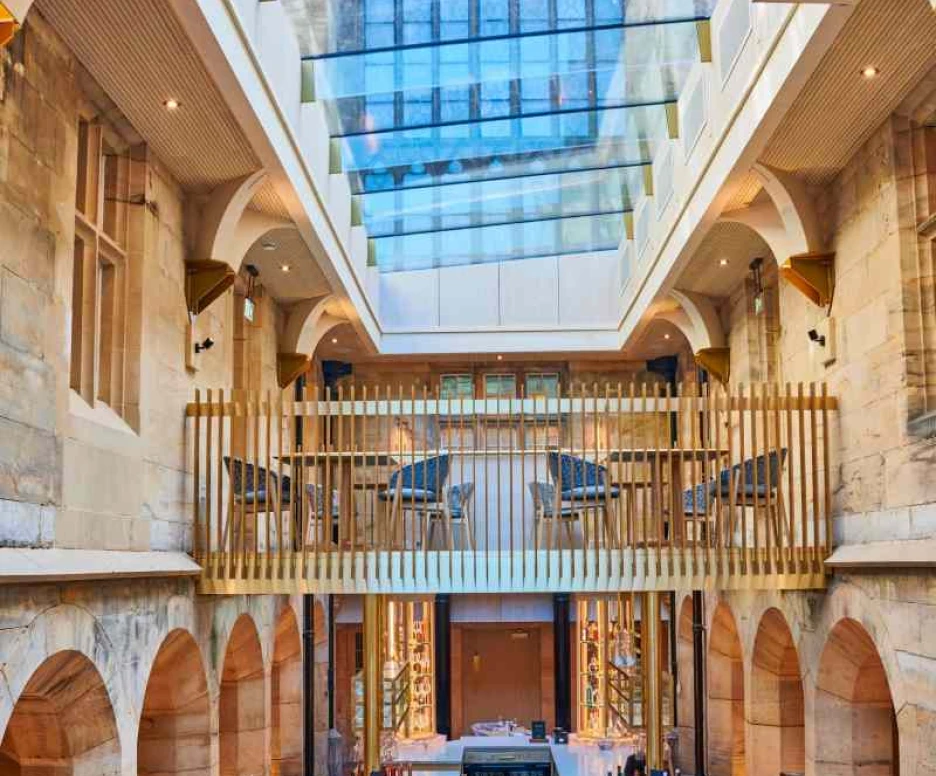 A modern restaurant interior featuring high ceilings, large glass windows, and an open layout. The space includes elegant wooden furnishings, archways, and a centrally located bar area with seating. Natural light floods the room, highlighting the architectural details.