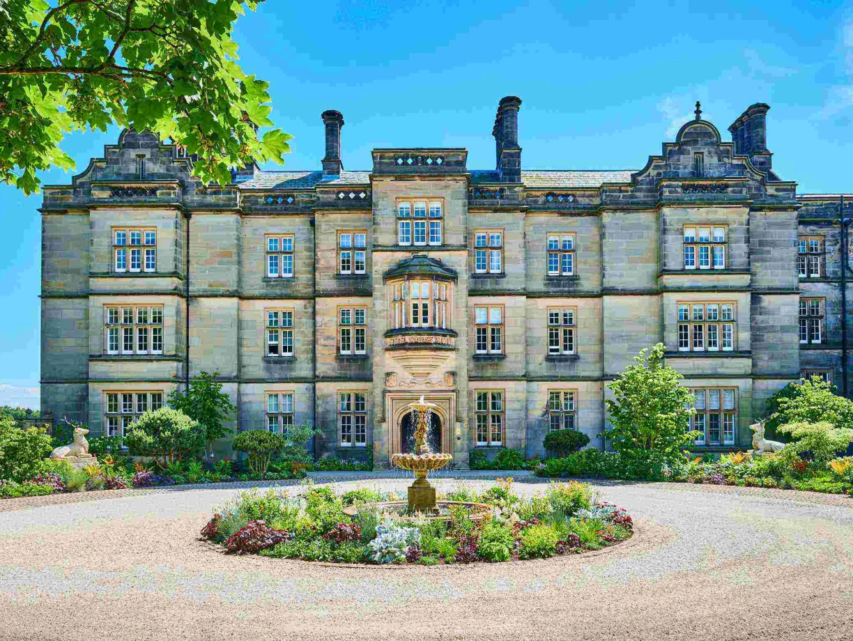 Historic stone building with intricate architecture, surrounded by colorful gardens and trees. A central fountain adds charm to the landscaped area in front. Bright blue sky enhances the scene.