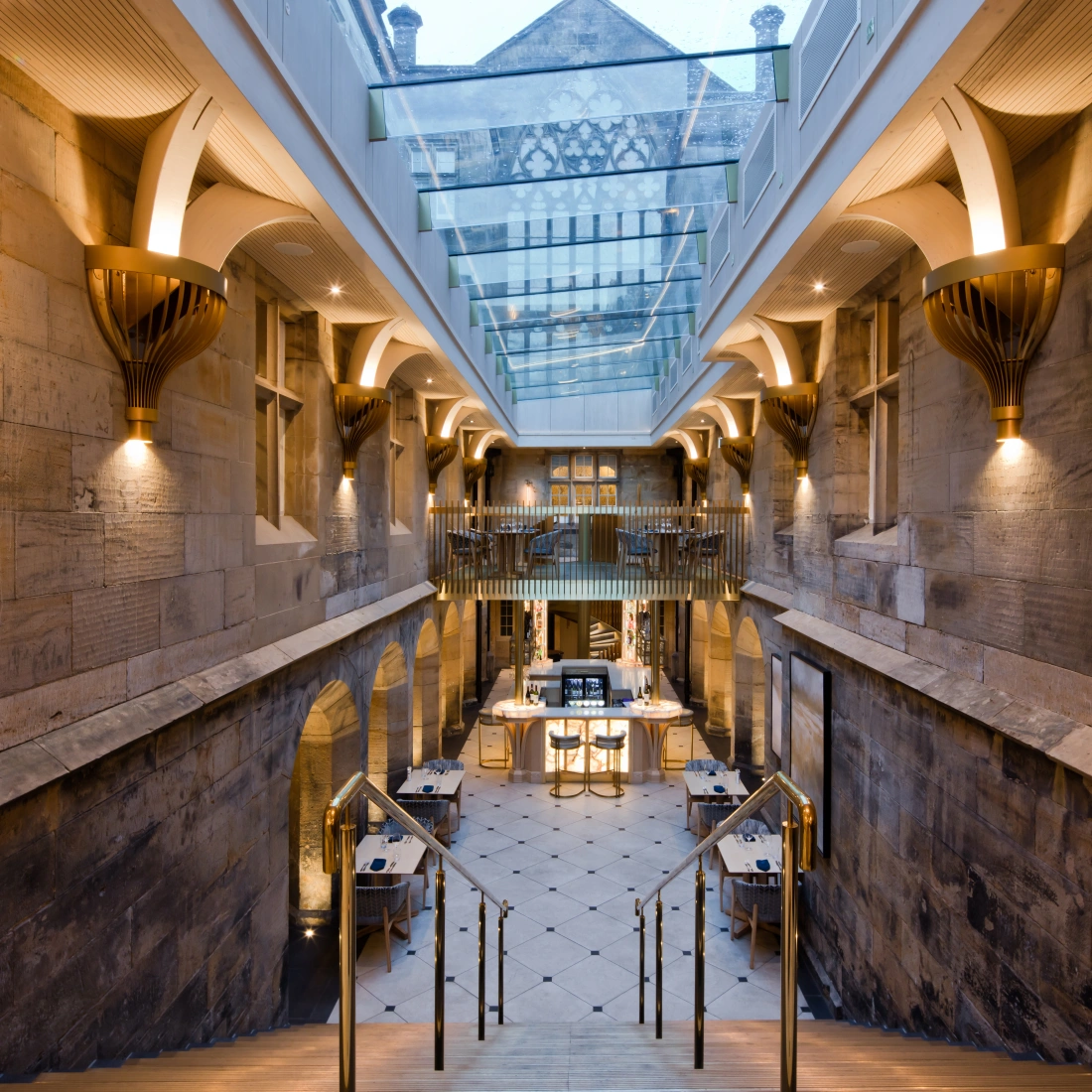 Interiores de un edificio con escaleras de piedra que conducen a un área abierta y elegante, iluminada por luces cálidas y un techo de cristal. Las paredes son de piedra y cuentan con detalles arquitectónicos distintivos.