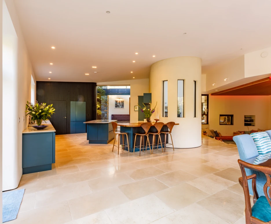 A spacious, modern kitchen and dining area featuring a large stone floor, light-colored walls, and contemporary furniture. The kitchen has dark cabinetry and a central island, while the dining space includes wooden chairs and a table. Natural light floods in through large windows.