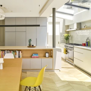 A modern kitchen featuring a light wooden dining table with two candles, a yellow chair, and a sleek island with storage. The kitchen has gray cabinetry, a white countertop, and stainless steel appliances, with a large window letting in natural light.