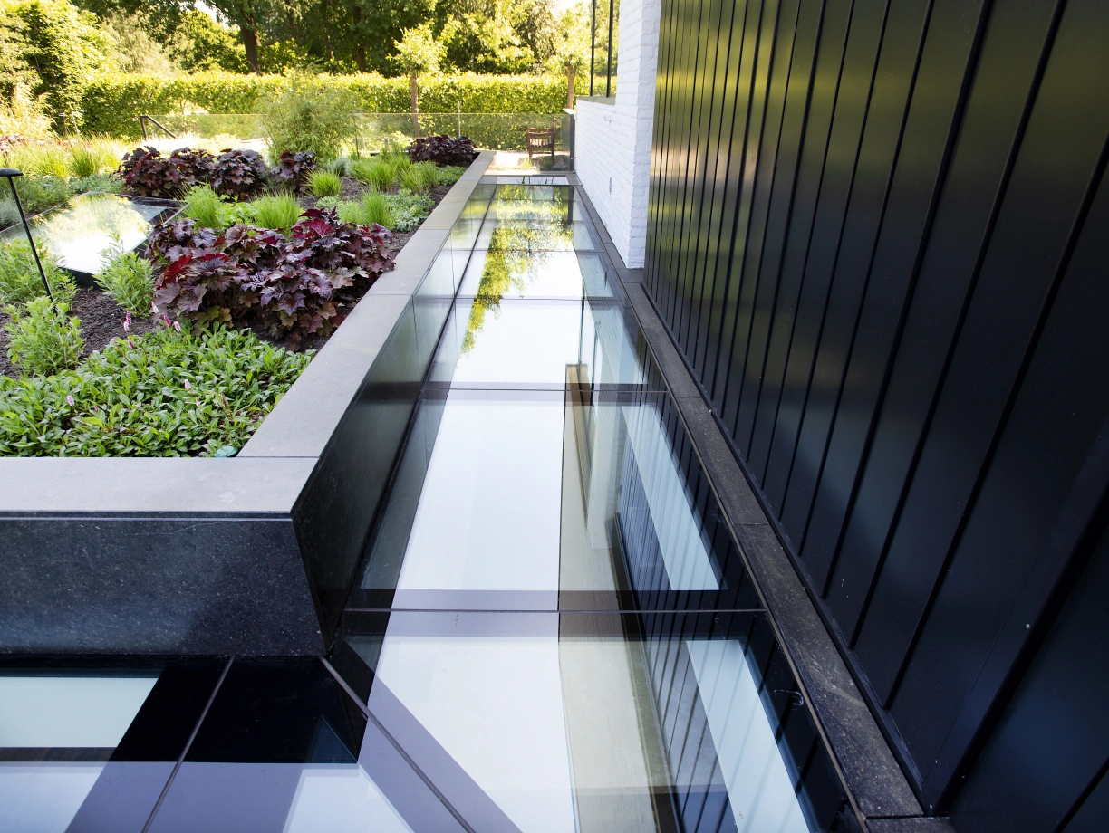Un chemin en verre transparent borde un jardin verdoyant avec des plantes et des feuillages colorés. Les murs noirs créent un contraste moderne avec la nature environnante.