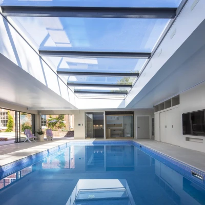Interior elegante con una piscina iluminada por una claraboya. Las paredes son de color claro y hay áreas para relajarse alrededor de la piscina. Se observa un patio exterior a través de las grandes ventanas.