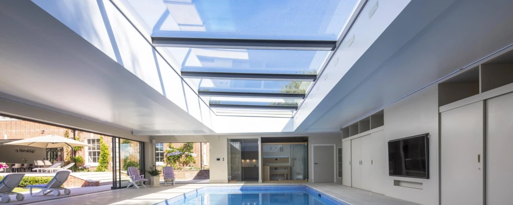 Intérieur d’une piscine avec un plafond en verre laissant passer la lumière naturelle. Des murs modernes et des portes coulissantes s’intègrent parfaitement au design. Des chaises longues sont disposées près de la piscine.