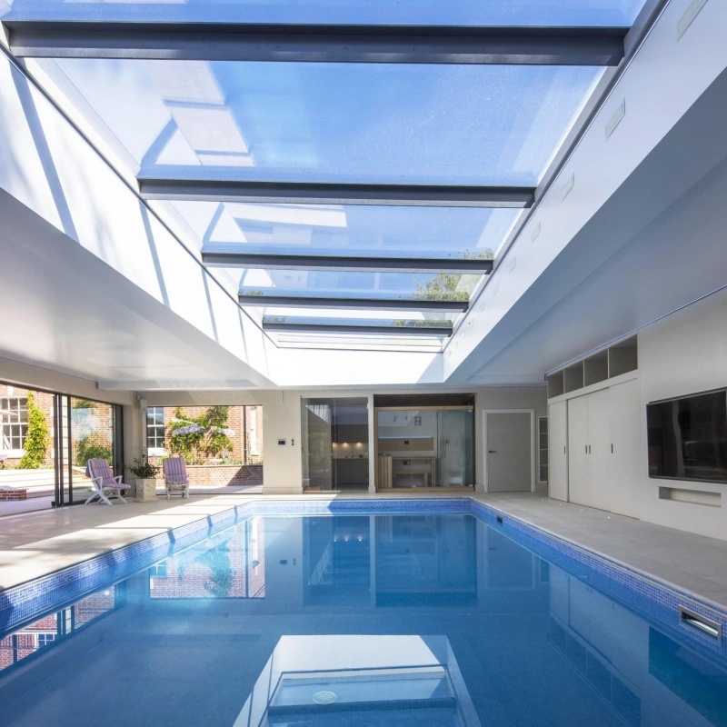 Intérieur d’une piscine avec un plafond en verre laissant passer la lumière naturelle. Des murs modernes et des portes coulissantes s’intègrent parfaitement au design. Des chaises longues sont disposées près de la piscine.