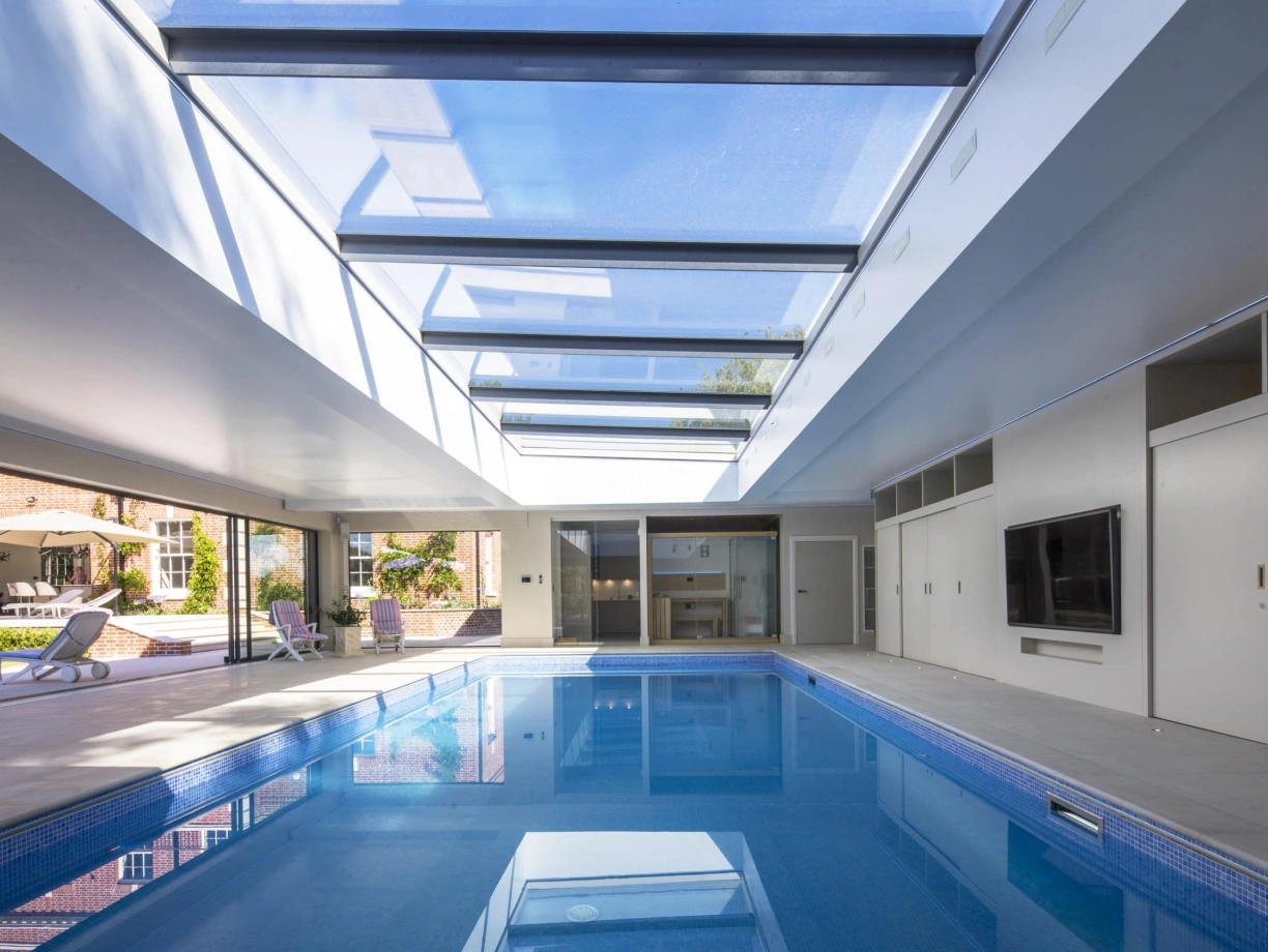 An indoor swimming pool with a sleek design, featuring a glass ceiling that allows natural light to flood the space. Modern lounge chairs are positioned around the pool, and large sliding doors lead to an outdoor area.