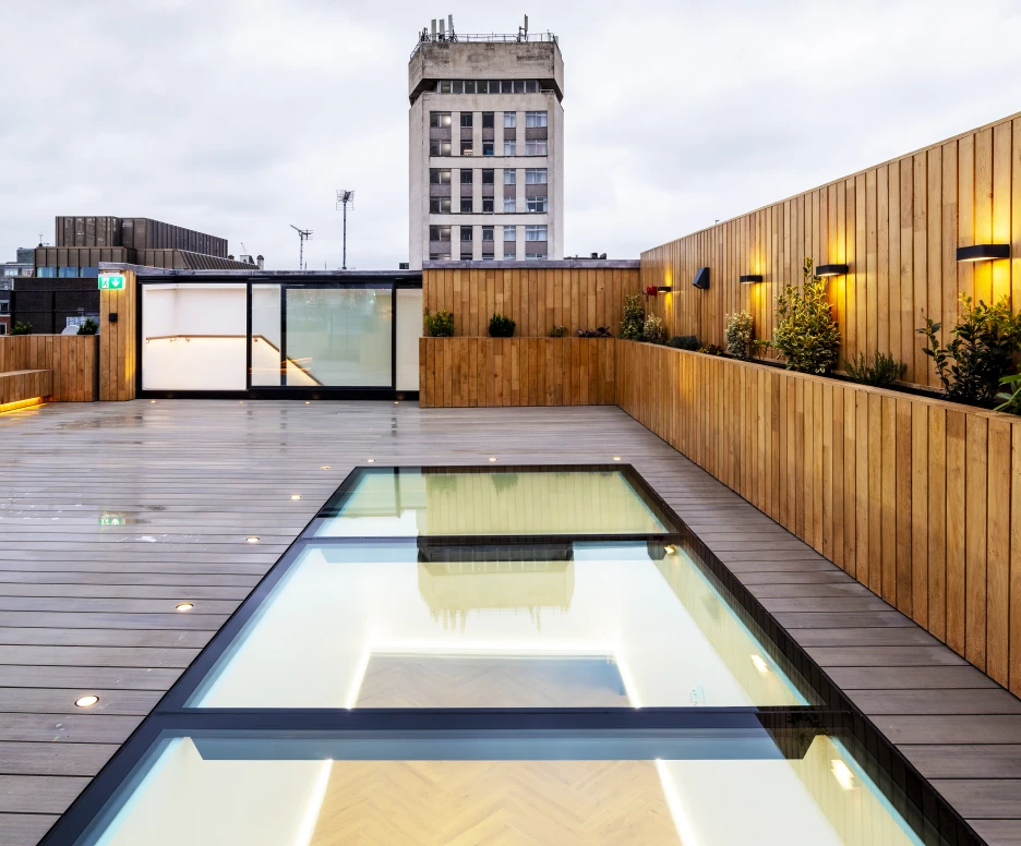 A modern rooftop with a wooden deck and a glass-lined swimming pool reflecting the overcast sky. A tall building stands in the background, adding to the urban atmosphere. Soft lighting accents the space, creating a warm ambiance.