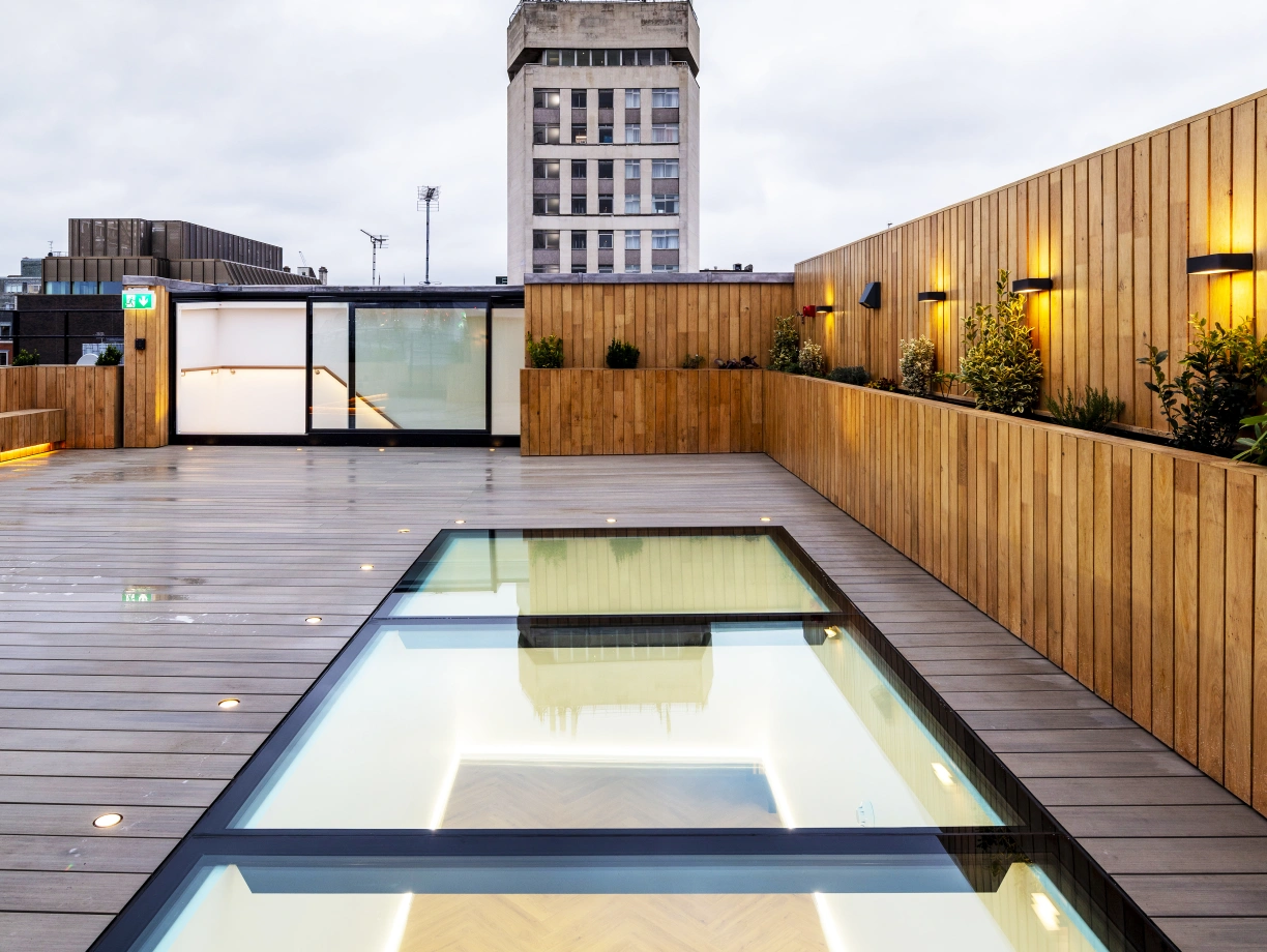 A modern rooftop terrace featuring a glass-bordered swimming pool reflecting the sky, surrounded by wooden decking and illuminated by soft lighting. In the background, a tall building with a classic facade is visible against a cloudy sky.