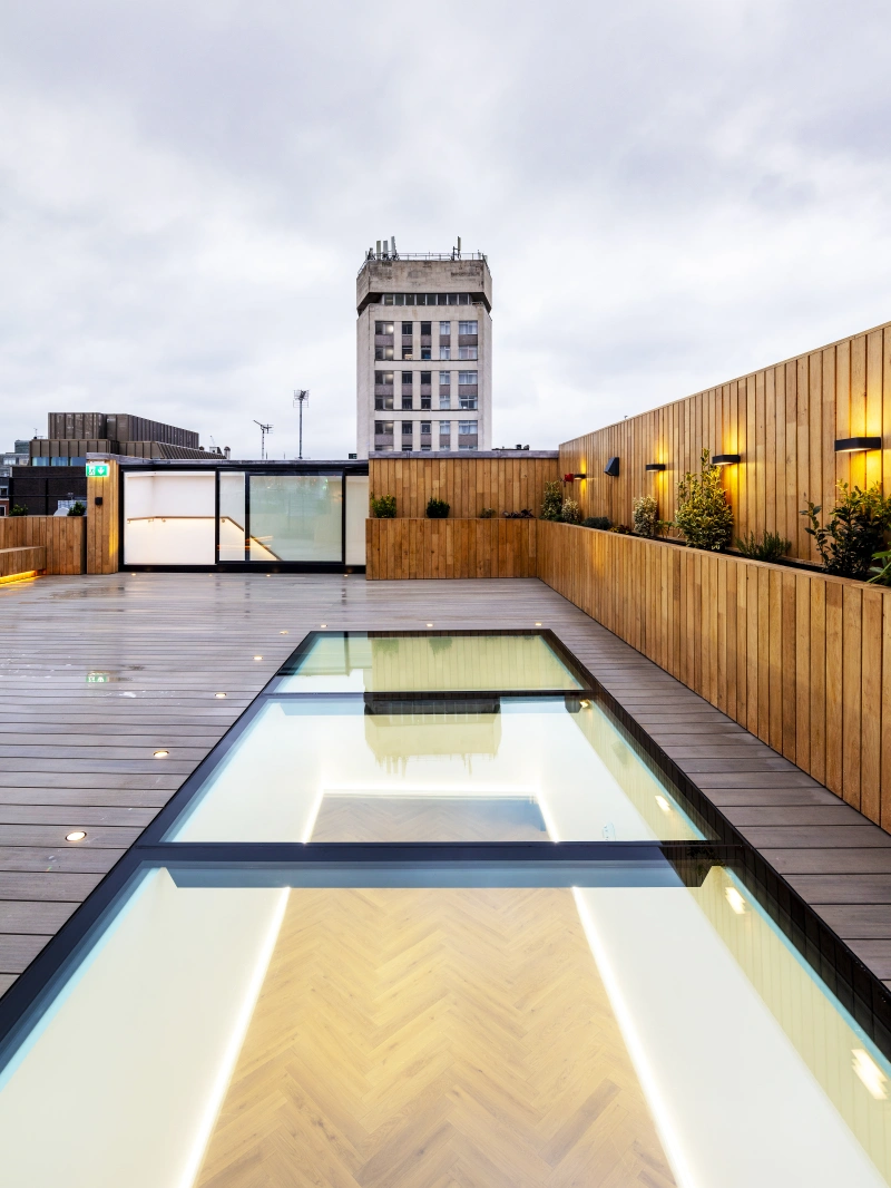 Une terrasse moderne avec un sol en bois, une piscine transparente et un bâtiment en arrière-plan sous un ciel nuageux. Des lampes éclairent la scène, créant une ambiance chaleureuse.