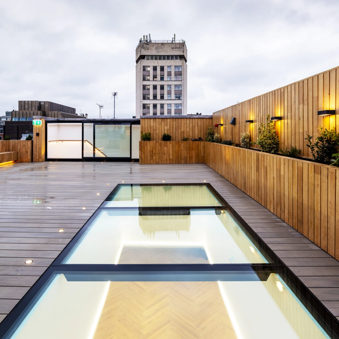 Une terrasse moderne avec un sol en bois, une piscine transparente et un bâtiment en arrière-plan sous un ciel nuageux. Des lampes éclairent la scène, créant une ambiance chaleureuse.