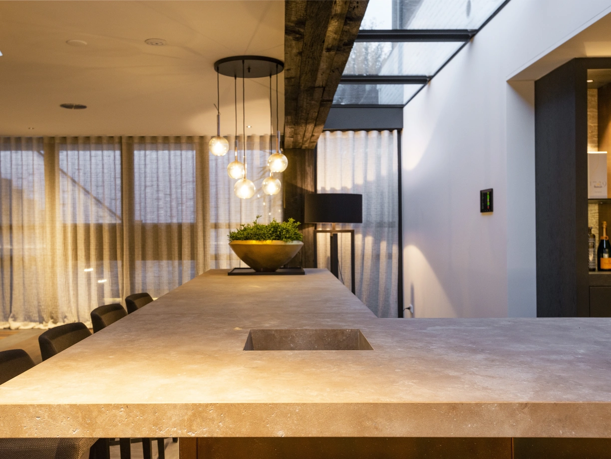 A modern kitchen with a large stone countertop featuring a sink cutout. Pendant lights hang above the counter, illuminating a planter with greenery. Floor-to-ceiling windows are partially covered by sheer curtains, and wooden beams accent the ceiling.