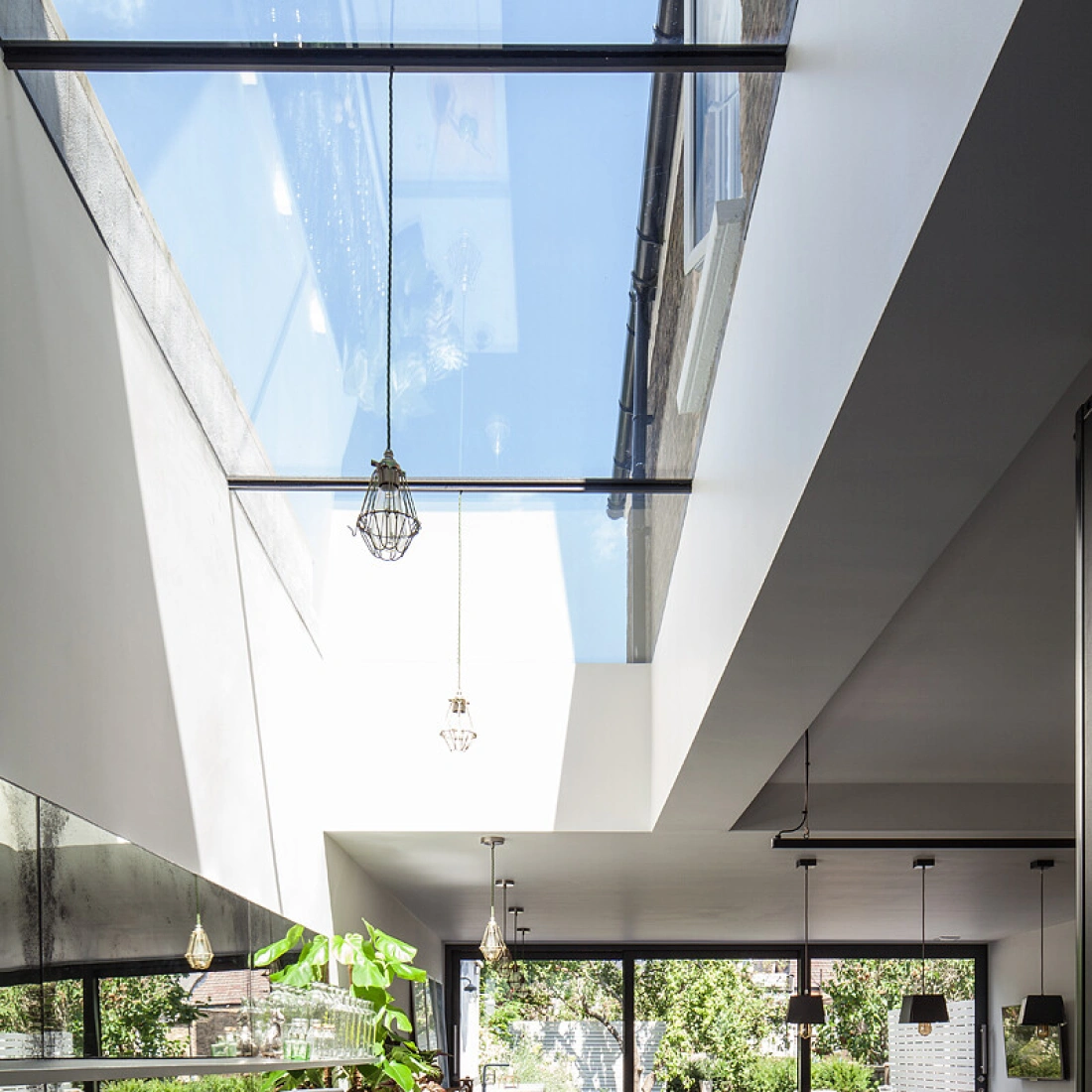 A modern kitchen featuring sleek black cabinetry, a concrete countertop, and large glass windows that let in natural light. The open layout includes a dining area with green plants and a view of a garden outside. Stylish pendant lights hang from the ceiling.