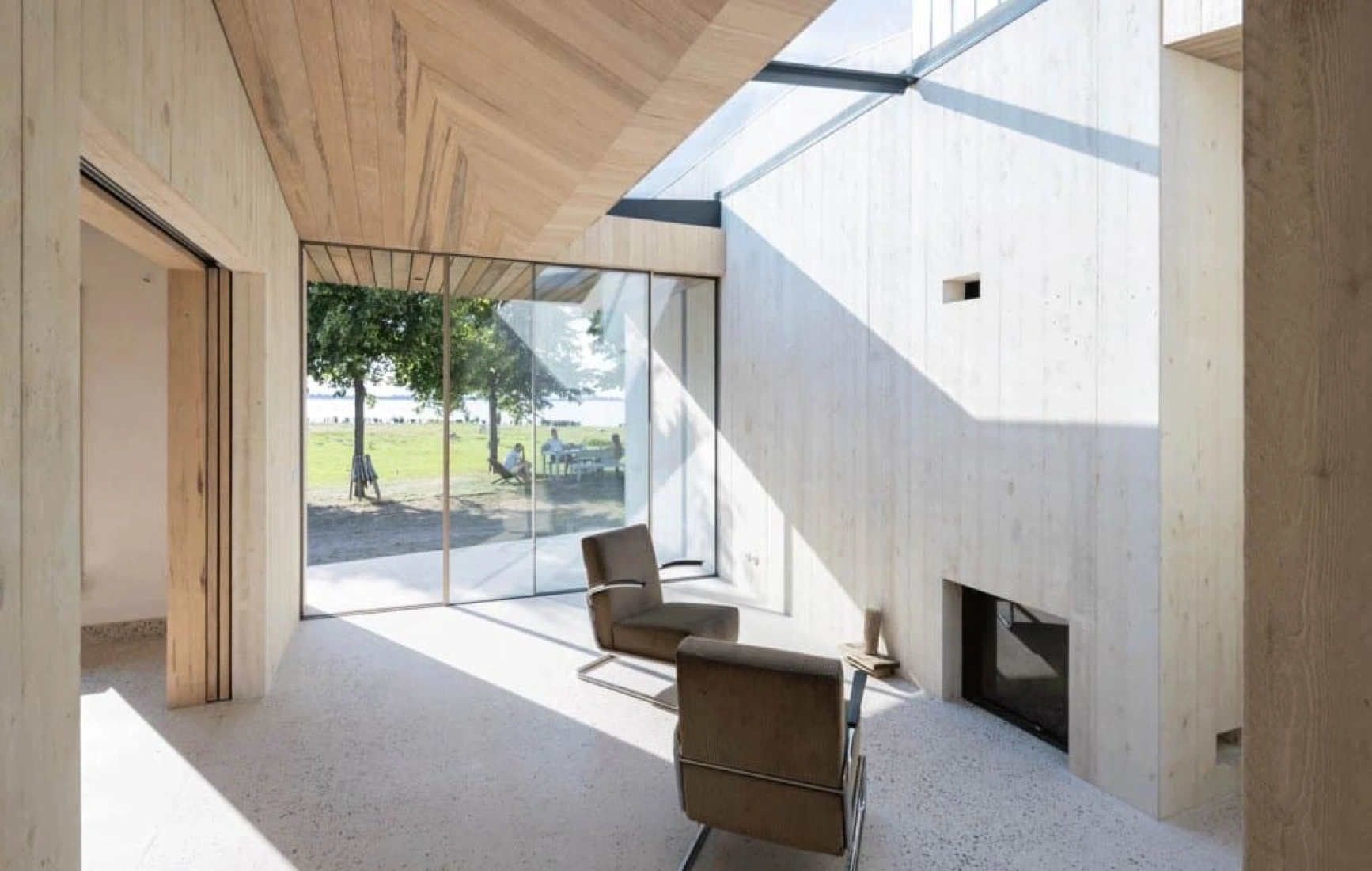 Moderne Architektur mit Holzverkleidung und großen Fenstern. Innenraum mit einem gemütlichen Stuhl und einem Kamin. Blick nach draußen auf eine grüne Wiese und Bäume. Tageslicht fällt durch die Fenster und schafft eine helle Atmosphäre.