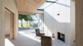 Moderne Architektur mit Holzverkleidung und großen Fenstern. Innenraum mit einem gemütlichen Stuhl und einem Kamin. Blick nach draußen auf eine grüne Wiese und Bäume. Tageslicht fällt durch die Fenster und schafft eine helle Atmosphäre.