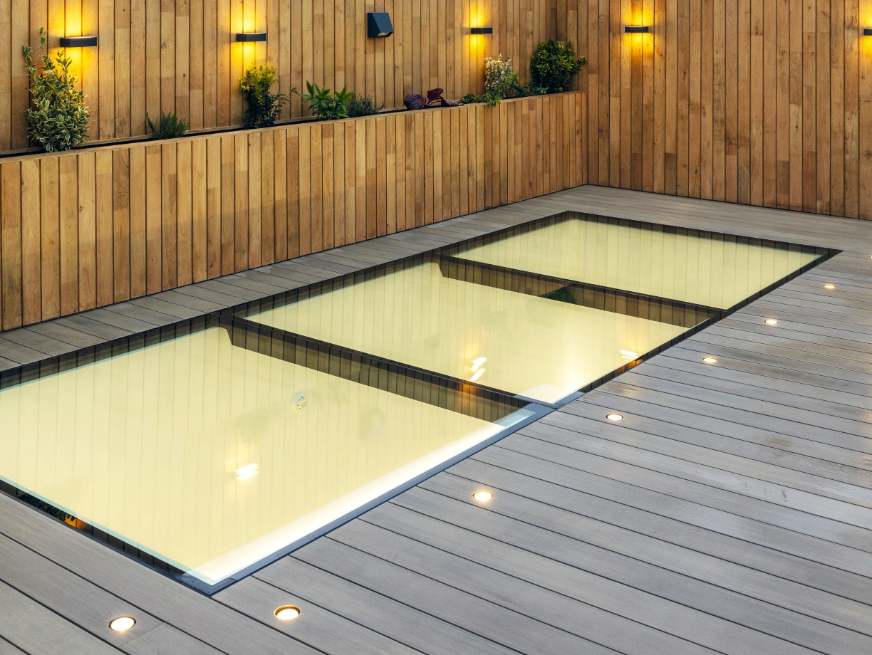 A sleek rooftop area featuring a modern, glass-walled pool surrounded by wooden decking. The wooden fence is adorned with small wall-mounted lights and plants. A cloudy sky can be seen in the background, along with a glimpse of a Ferris wheel.