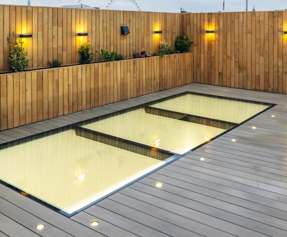 A sleek rooftop area featuring a modern, glass-walled pool surrounded by wooden decking. The wooden fence is adorned with small wall-mounted lights and plants. A cloudy sky can be seen in the background, along with a glimpse of a Ferris wheel.