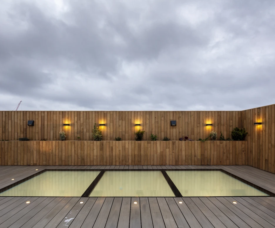 A modern rooftop space featuring a rectangular pool set within a wooden deck, surrounded by a wooden wall adorned with plants and wall-mounted lights. The sky is overcast with clouds.