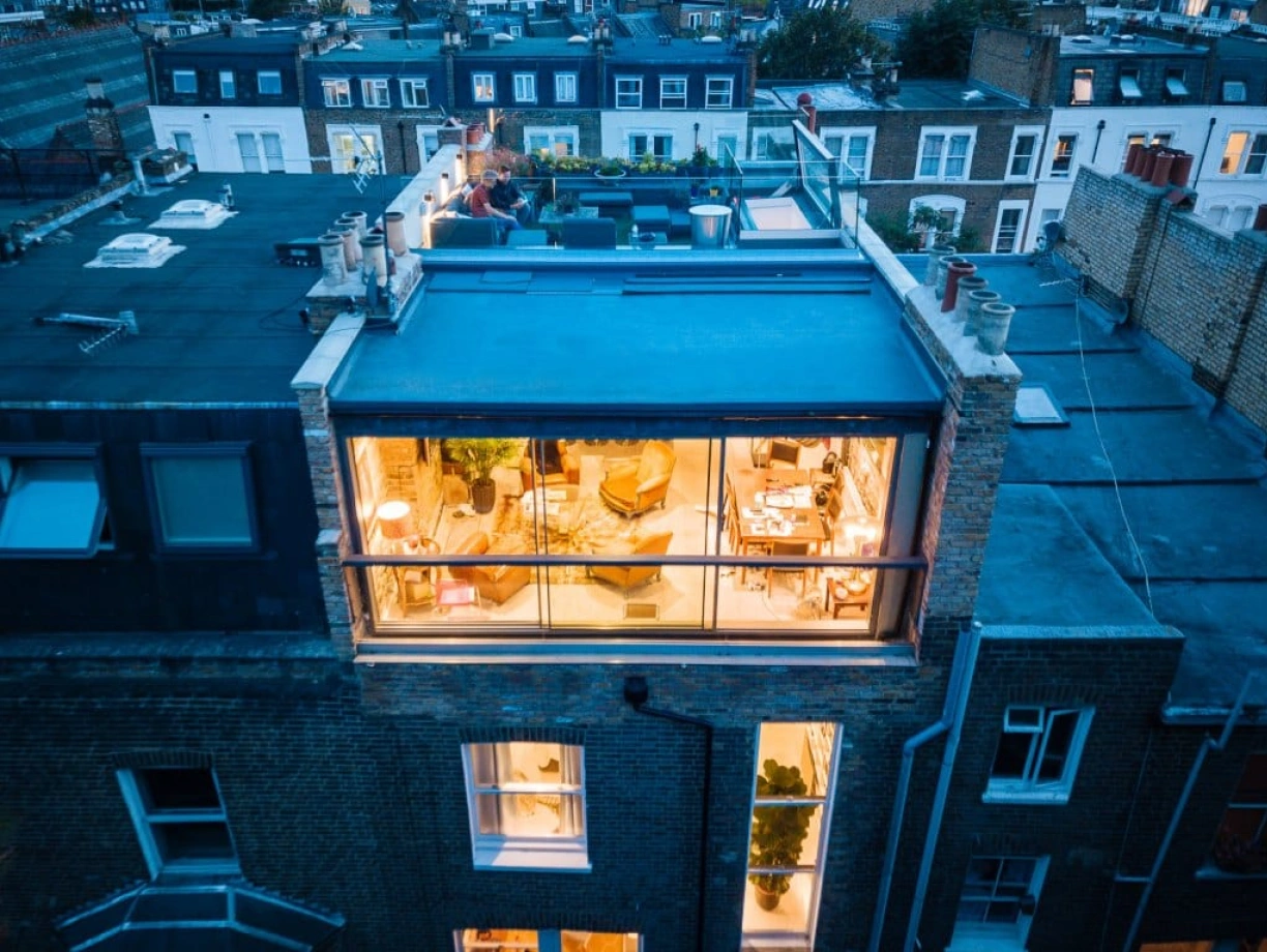A modern rooftop terrace with a glass-walled lounge area, featuring cozy furniture and decorative plants. Nearby rooftops of city buildings are visible in the background, illuminated by evening lights.