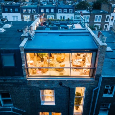 A modern rooftop terrace with a glass-walled lounge area, featuring cozy furniture and decorative plants. Nearby rooftops of city buildings are visible in the background, illuminated by evening lights.