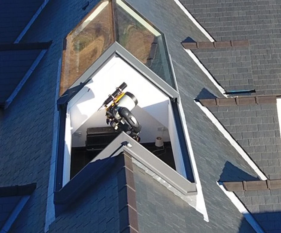 A sleek, modern rooftop with a distinctive angular skylight, featuring a small telescope and a viewing platform inside. The surrounding roof is covered with dark shingles, enhancing the contemporary design.