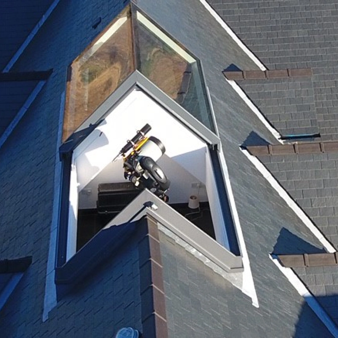 A sleek, modern rooftop with a distinctive angular skylight, featuring a small telescope and a viewing platform inside. The surrounding roof is covered with dark shingles, enhancing the contemporary design.