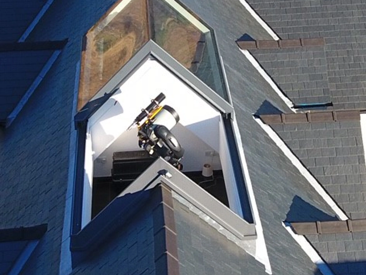 A sleek, modern rooftop with a distinctive angular skylight, featuring a small telescope and a viewing platform inside. The surrounding roof is covered with dark shingles, enhancing the contemporary design.