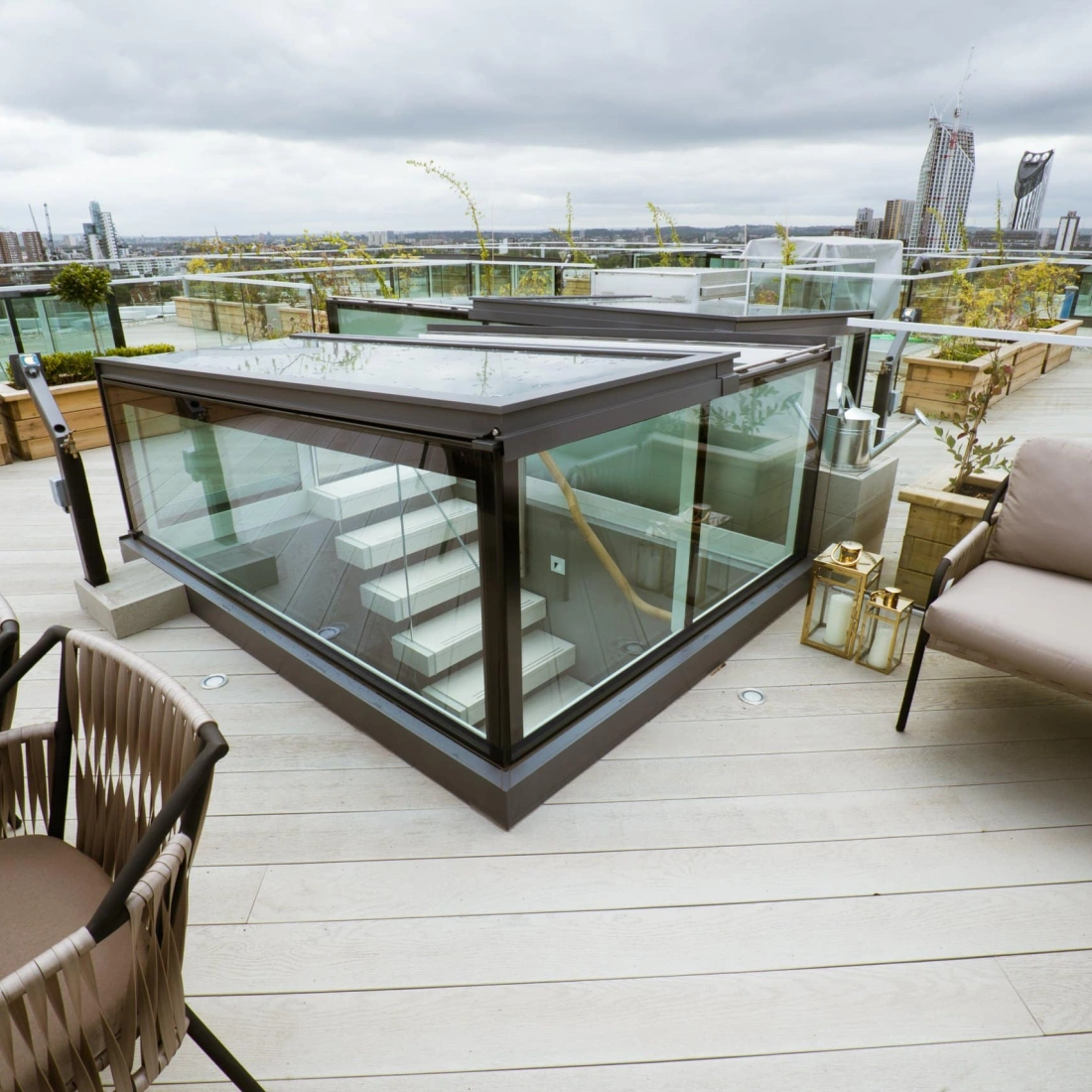 A modern rooftop terrace features a glass enclosure and a staircase leading down. Stylish outdoor seating with a table is provided, along with planters and greenery. The skyline and tall buildings are visible in the background under a cloudy sky.