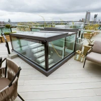 Terraza moderna con barandillas de vidrio, escaleras y muebles de exterior. Se observa una vista urbana con edificios al fondo. El cielo está nublado.