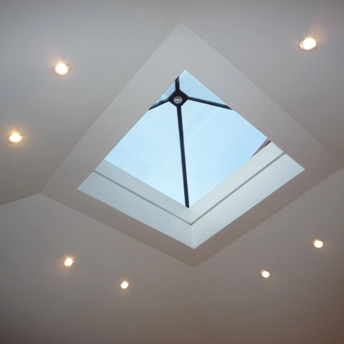A modern ceiling with recessed lighting and a square skylight that shows a clear blue sky.