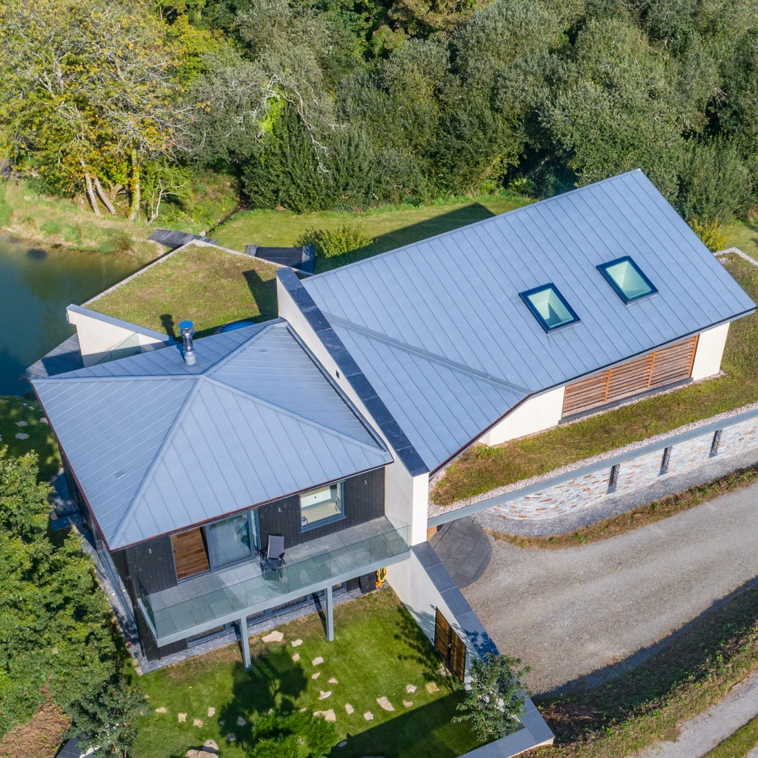 Moderne Architektur eines Hauses mit einem schrägen Dach, umgeben von Bäumen und einem kleinen Teich. Eine geschwungene Auffahrt führt zum Eingang des Hauses.